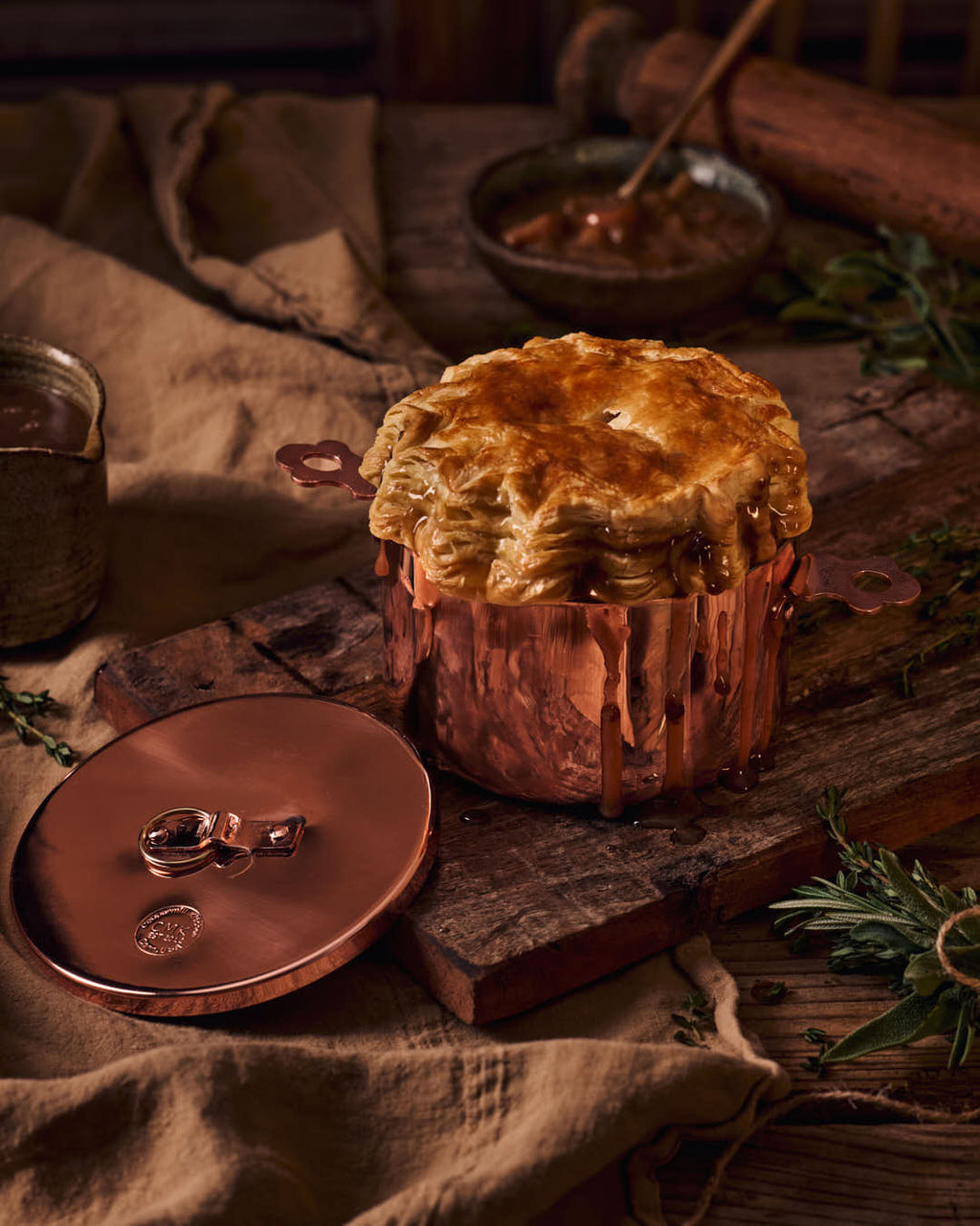 Copper pot with a pie on a rustic wooden surface with herbs and a candle.