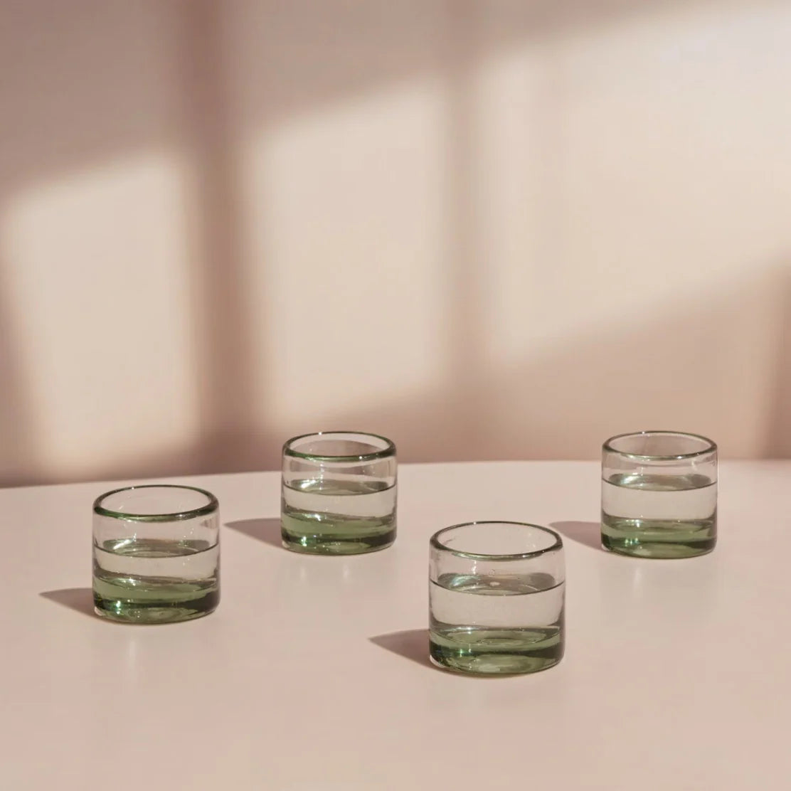 Four green glass tumblers on a light pink surface with a soft pink background