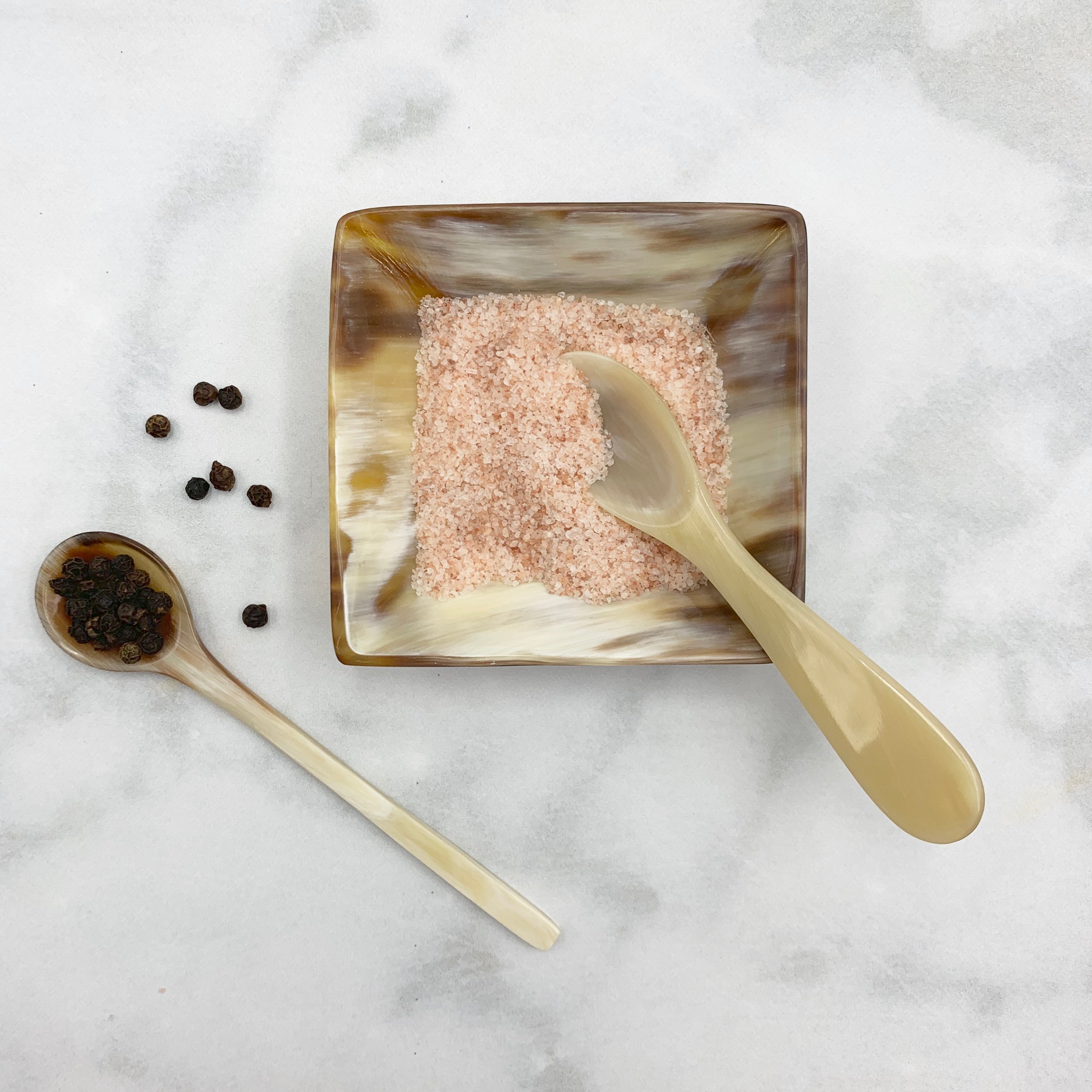 Square bowl with pink salt, gold spoon, and black peppercorns on a marble surface