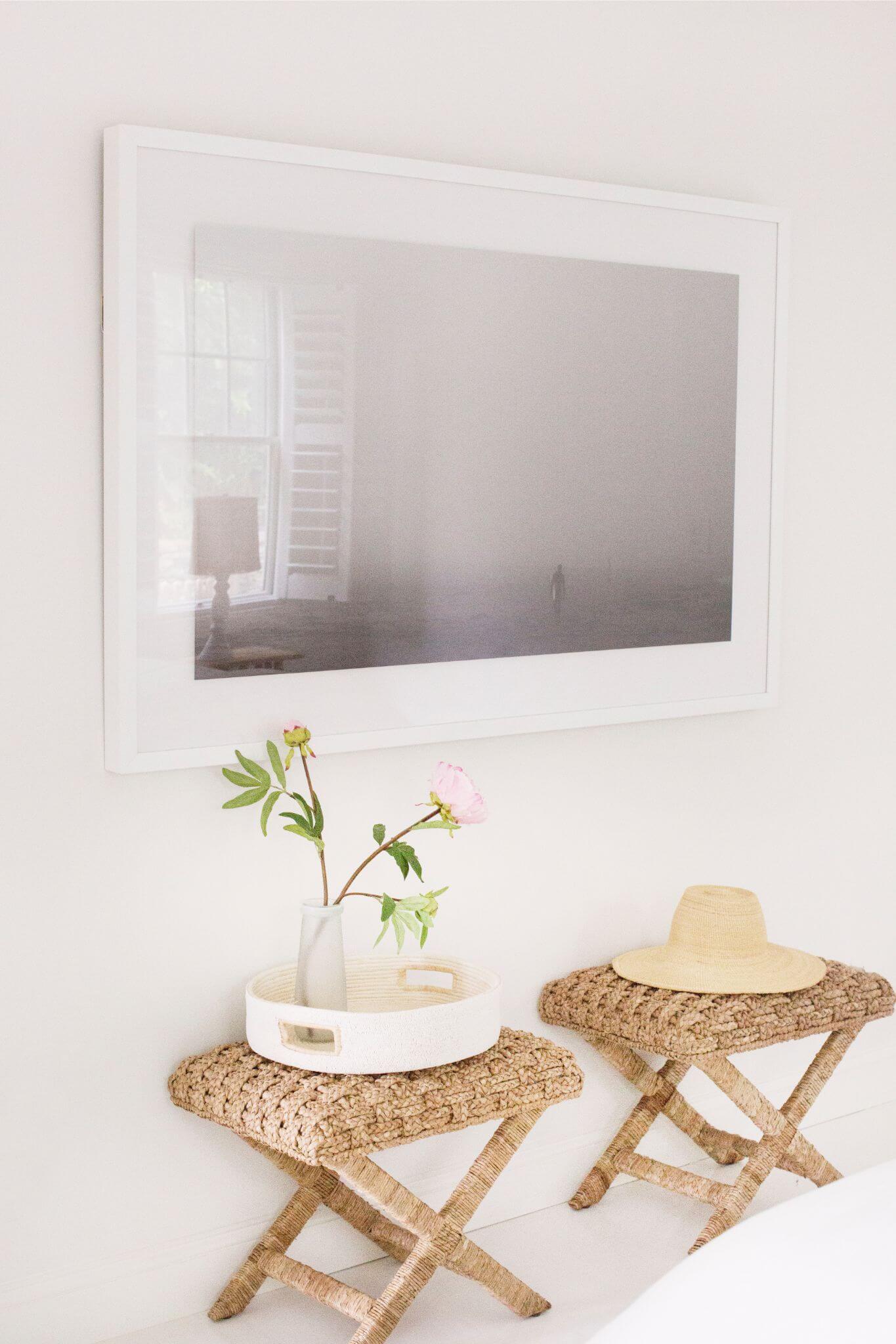 Two woven stools with a tray and hat against a white wall with a mirror.