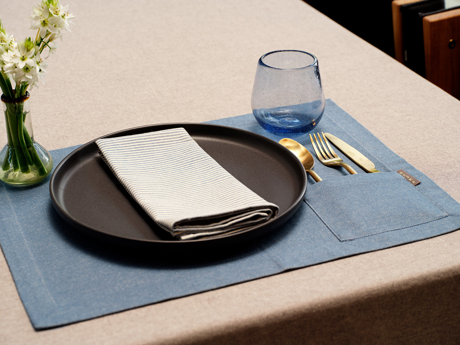 Dining table setting with black plate, folded napkin, blue glass, silverware, and blue placemat.