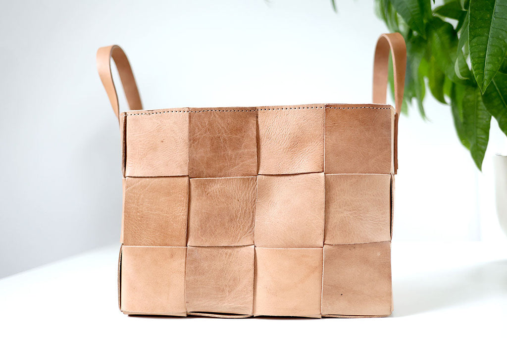 Woven leather basket with handles on a white surface with a plant in the background