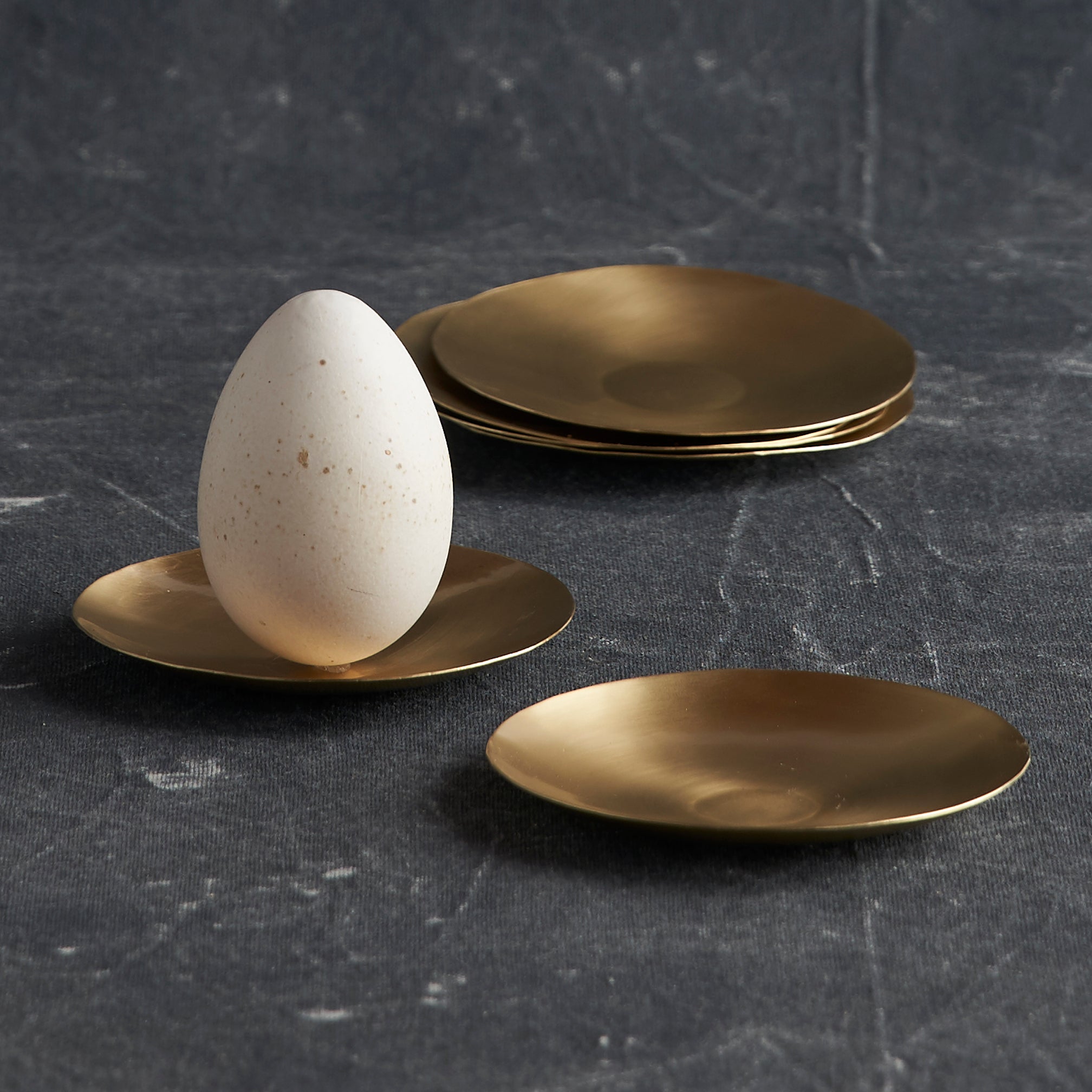 White egg on a gold plate with additional gold plates on a dark textured surface