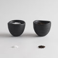 Two black ceramic bowls with salt and pepper on a light gray background