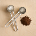 Two metal coffee scoops on a beige surface with ground coffee.