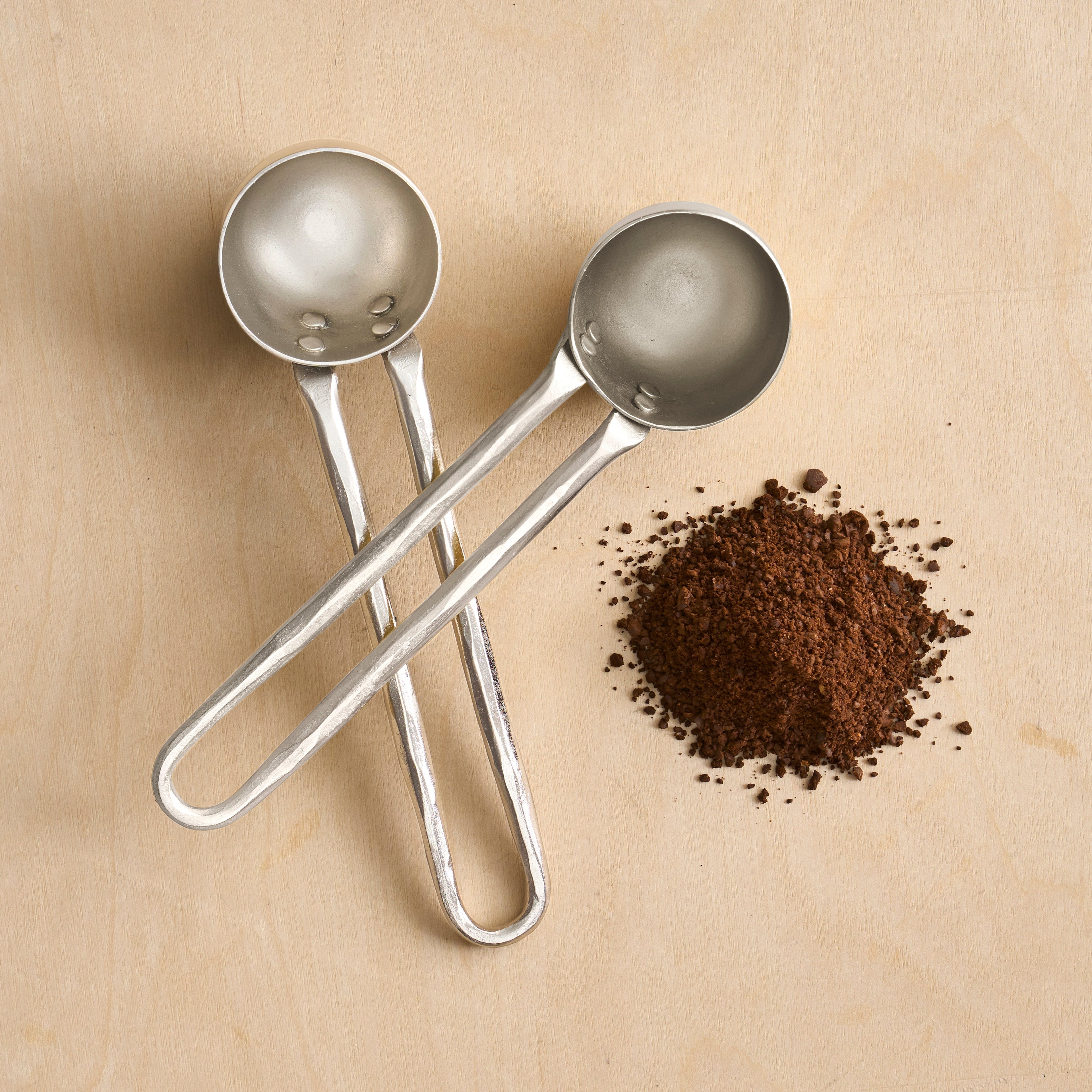 Two metal coffee scoops on a beige surface with ground coffee.