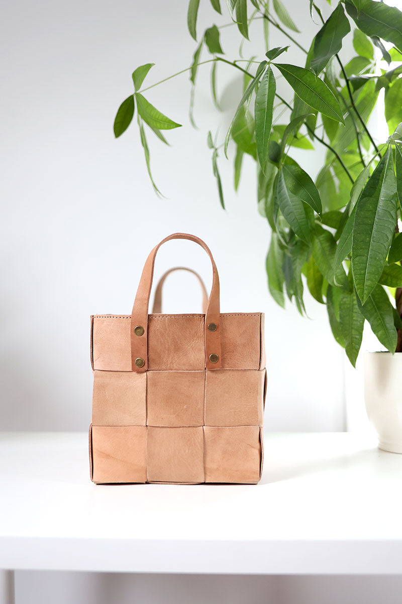 Brown woven bag on a white surface with green leaves in the background