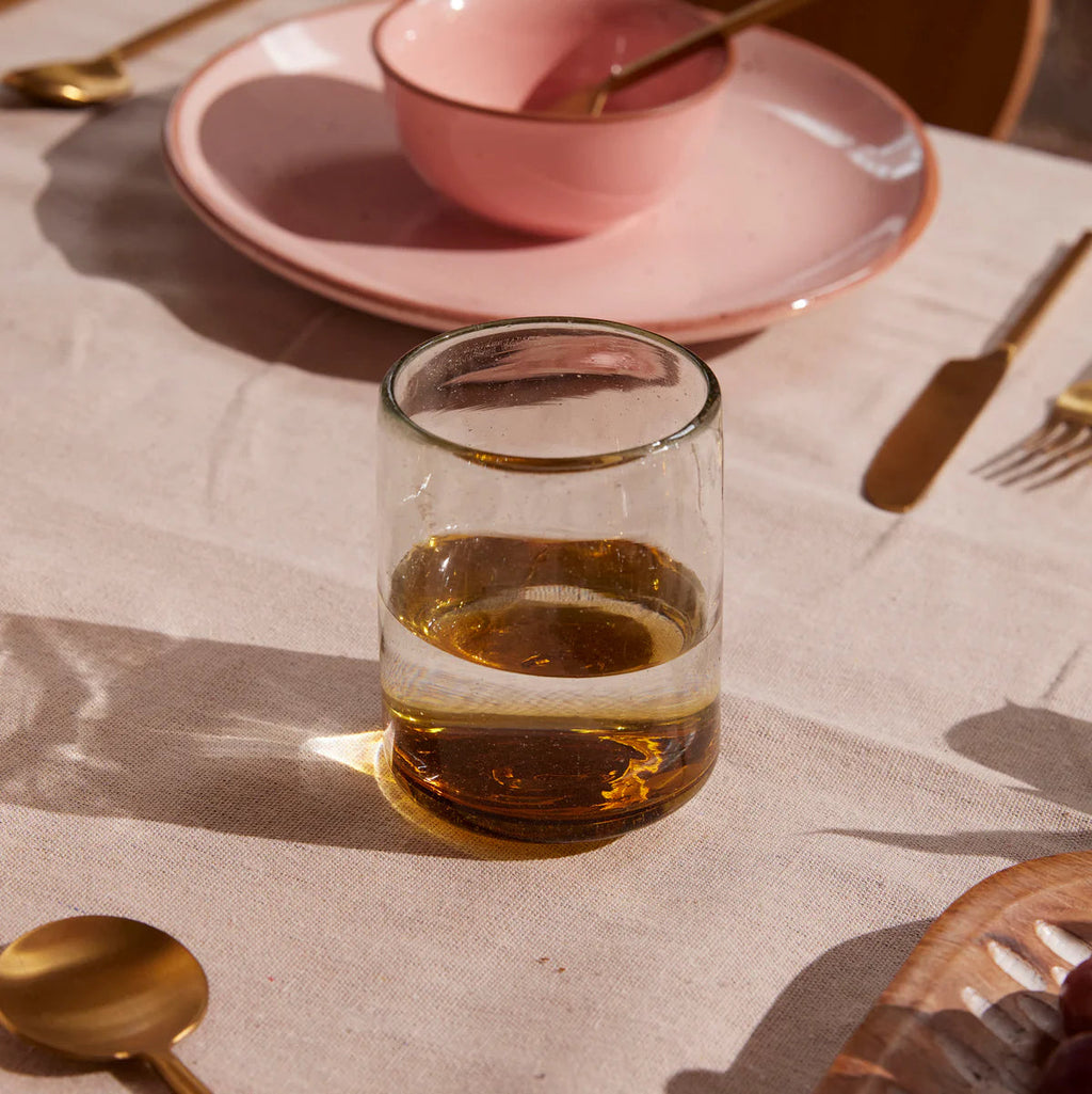 Clear glass with amber liquid on a tablecloth with cutlery and pink plates.