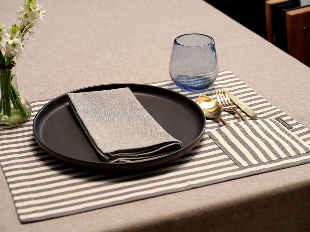 Dining table setting with black plate, gray napkin, blue glass, and striped placemat.