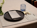 Dining table setting with black plate, gray napkin, blue glass, and striped placemat.