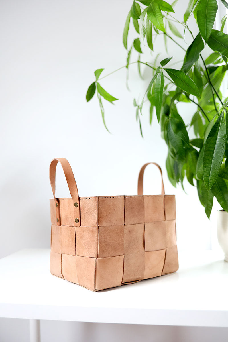 Brown woven basket with leather handles on a white surface with a green plant in the background